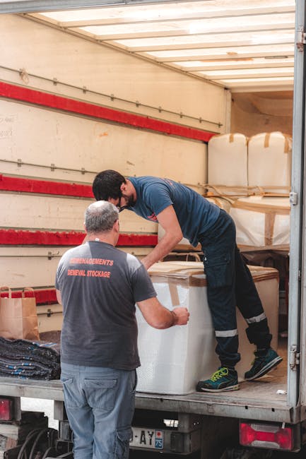 A man wearing a black cap, white polo shirt, and jeans is kneeling on the pavement outside a property, closely inspecting and marking cardboard boxes used for home relocation. The boxes vary in size a